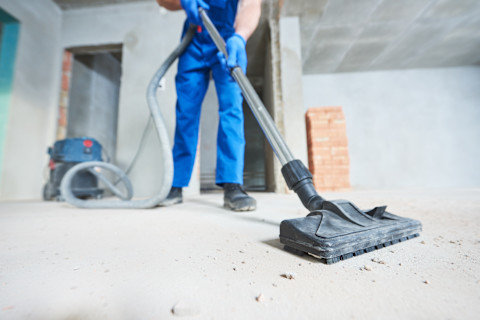 A worker in blue work clothes vacuums a recently constructed, unfinished room. Dust particles are on the ground.