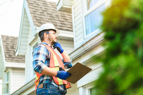 A worker, wearing safety gear, at the top of a ladder, holding a clipboard and looking at a rooftop while thinking.
