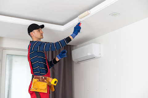 A worker with overalls and a toolbelt stands on a ladder using a roller to paint the ceiling.