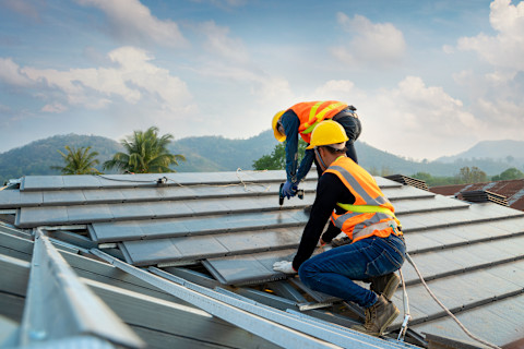 Two construction workers wear safety gear, including hard hats and harnesses, as they install concrete roof tile.