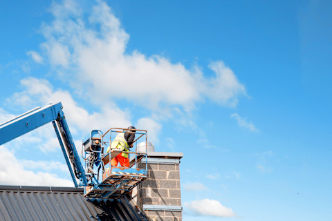 This image shows tradespeople in the basket of a crane fixing the chimney on a rooftop. 