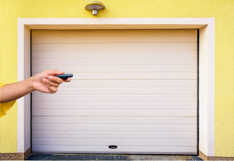 Frontal shot of a white garage door in a yellow house, with a hand holding a remote coming in from the left side of the frame.
