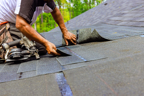 A roofing pro installs new grey shingles on a roof. A nail gun is at his side.
