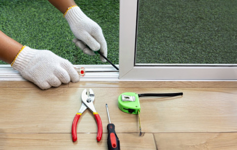 A worker fixes a derailed sliding glass door. Closeup on hands with tools visible on the ground.