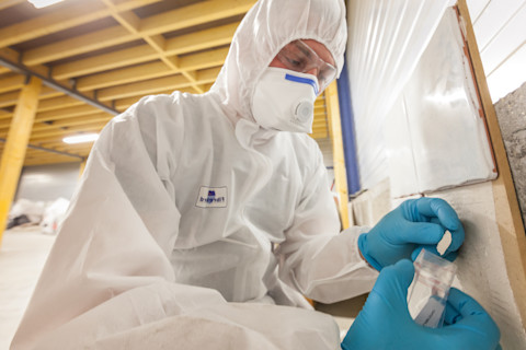 An asbestos surveyor in protective gear collects a sample of building material in a building under construction.