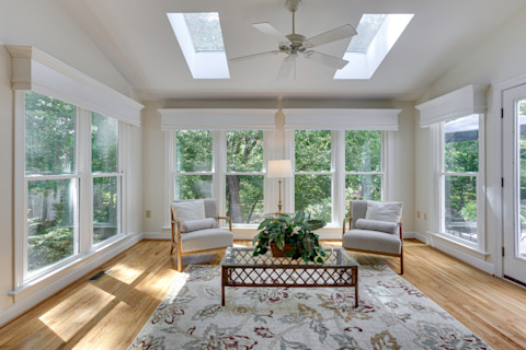 Bright sunroom with large windows and skylights, two cushioned chairs, a glass coffee table with a plant, a rug over the wooden floor, and an inviting atmosphere.