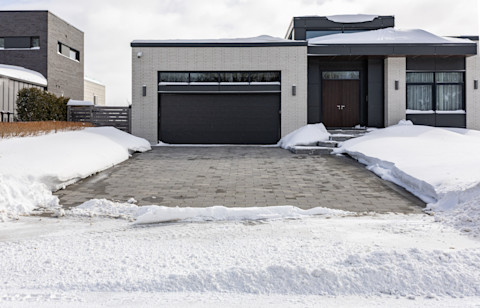 The image shows a heated driveway in front of a single storey home cleared from snow after a storm. The surrounding ground is covered with thick white snow. 