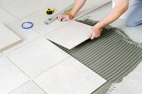 This image shows a tile fitter kneeling down to carefully lay a ceramic tile on grey cement, ensuring proper alignment and smooth installation. To the side of them, there is a tape measure and spirit level. 