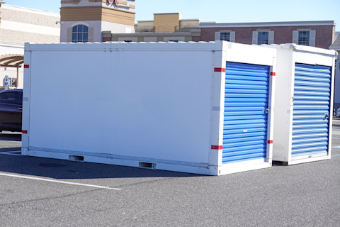 A white storage unit with bright blue rolling doors sits in a parking lot on a sunny day. A building with brick accents is in the background.
