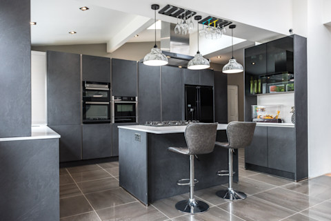 A large, minimalist style black and grey kitchen, with a breakfast and two stools in the centre beneath three hanging, grey marbled lights. There are several built-in cupboards around the perimeter, with an oven, microwave, and fridge built in to the structure. 