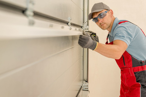 Side angle of a worker in overalls, cap, gloves and safety goggles installs a new residential garage door.