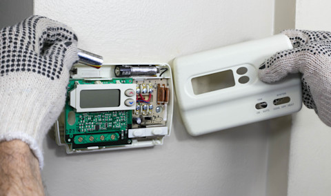 Wall-mounted thermostat installation with visible circuit board, allowing control of heating and cooling systems. The technician connects the wiring and checks internal components. This type of regulation optimizes thermal comfort while reducing energy consumption.