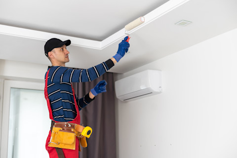 A worker with overalls and a toolbelt stands on a ladder using a roller to paint the ceiling.