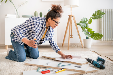 This image shows a woman kneeling down surrounded by three planks of white wood, a drill and other tools. She has a screwdriver in her right hand and is ready to assemble the pieces of wood. She is in a bright, modern living room and wearing casual clothes.