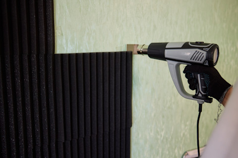  A technician uses a hot glue gun to attach foam panels to a wall. Only the technician’s hands are visible.