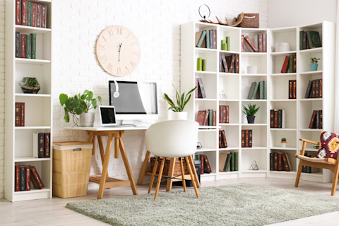 Modern, white office with desk, computer, and chair in the centre pushed against a wall, underneath a large clock hanging on the wall. Several bookcases border the rest of the wall beside the desk. A large grey rug is spread along the floor.