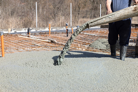 A worker guides the spout of a concrete mixer on a construction site as concrete pours out, forming a foundational slab.