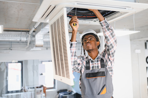 A technician in work attire cleans an outdoor air conditioning unit near a wooden wall.