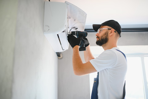 An HVAC technician uses a power drill to install an air conditioning unit on the wall.
