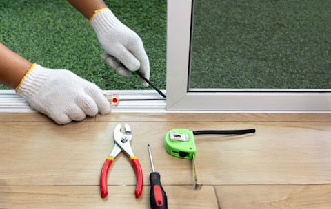 A worker fixes a derailed sliding glass door. Closeup on hands with tools visible on the ground.
