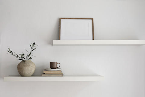 This image shows two white floating walls attached to a white wall. On one shelf, there is a picture frame, and on the other, there is a ornamental plant and a couple of books with a mug on top.