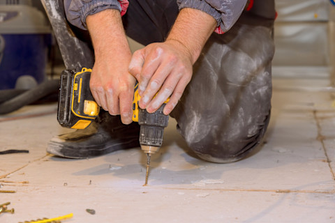A worker kneels on the floor as he uses a drill to repair a plywood subfloor. Closeup on hands.