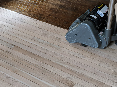 A worker kneeling on a hardwood floor pours wood filler from a bottle onto a damaged area of the floor during a refinishing project.