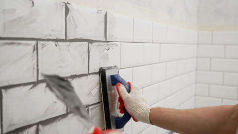 A tiler applies grout to a white tile backsplash, probably in a kitchen.