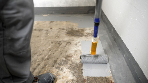 A worker applies a grey waterproofing membrane in a basement using a paint roller.