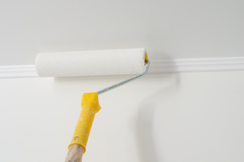 Closeup of a paint roller with a yellow handle applying white paint or primer to a ceiling.