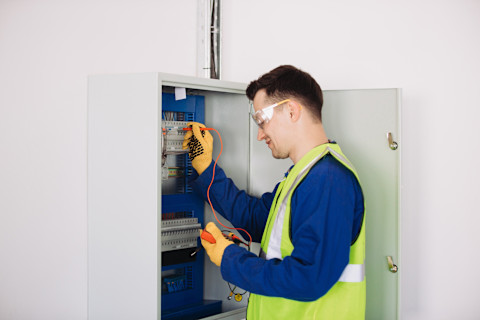 A worker wearing safety glasses and reflective vest checks a fuse box with a multimeter.