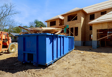 This image shows a blue dumpster container in the front yard of a house that is undergoing construction. The container is filled with scrap materials from the build.