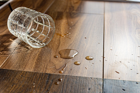 A glass of water tipped on its side next to some drops of water on a wooden laminate floor.