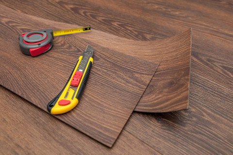 Some vinyl floor panels lay on some installed floor panels of the same design, with a measuring tape and box cutter on top.