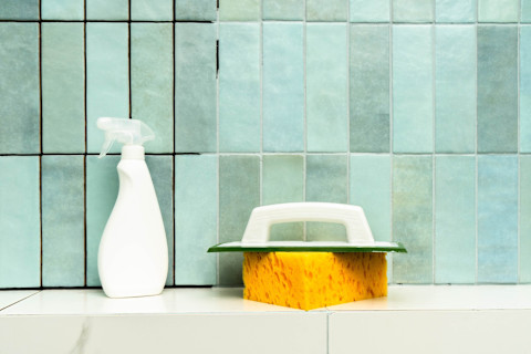 A spray bottle, sponge, and grout cleaner sit on the edge of a bathtub right next to the bathtub wall, with green and blue tile.