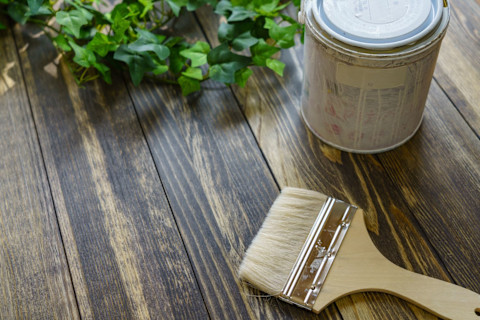 Closeup of a paintbrush and can of paint sitting on a treated wooden surface. Ivy is visible along the upper edge of the photo.
