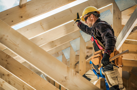 A construction worker wearing a yellow hard hat, safety glasses, and a tool belt inspects wooden roof framing on a new home build.