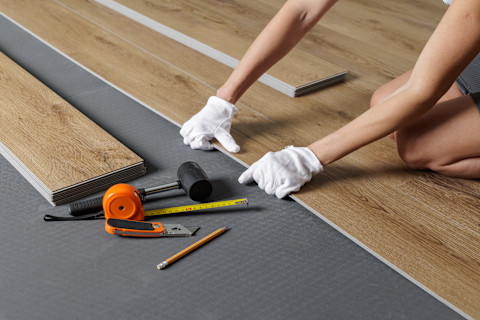 A worker kneels on the floor and adjusts a panel during a vinyl plank flooring installation. A jackknife, rubber mallet, measuring tape, box cutter and pencil are laying on the ground.