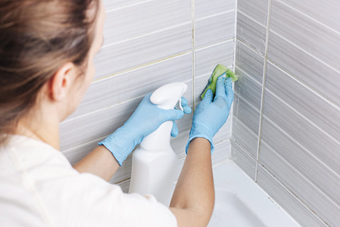 A cleaner wearing gloves uses a spray bottle and sponge to clean grout from the walls surrounding a bathtub.