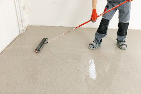 A worker wearing spiked shoes uses a spiked roller to smooth freshly poured epoxy across a concrete floor. Only the worker's legs and feet are visible.
