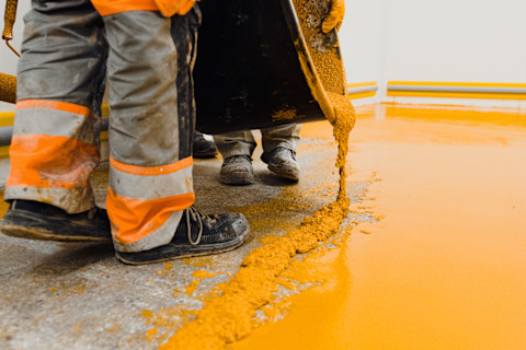 Two workers in industrial coveralls pour a yellow floor coating mixture from a scoop onto a concrete floor.