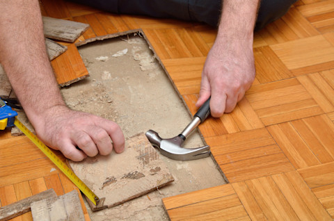 Closeup on the hands during a hardwood flooring project. Some panels have been taken out, so a concrete subfloor is visible, along with a hammer and measuring tape.