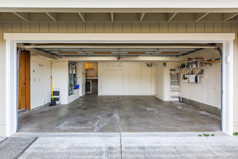 Empty two-car garage with an open door, showing a bare, stained concrete floor, a stepladder and ladder, a broom, and a wall-mounted tool shelf.