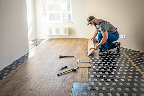 Wide angle of a worker installing flooring in a home. A few tools including a hammer and measuring tape are laying on the ground.