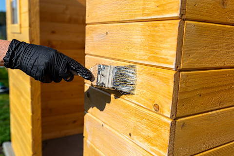 Closeup on a worker’s gloved hand applying finish to a wooden garden shed.