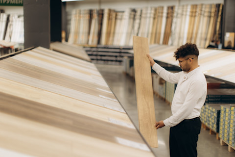 A man examines a wooden plank sample in a store. Several other samples lay in front of him on a shelf.