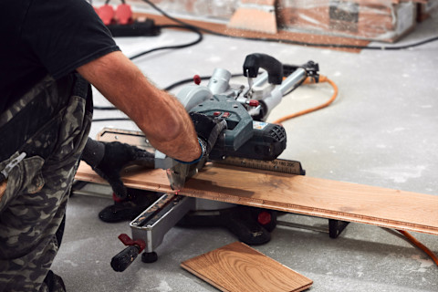 A worker uses a saw to cut laminate flooring on a worksite in a home.