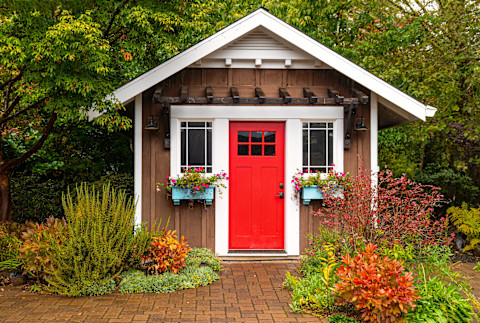 Frontal shot of a dark brown garden shed with a bright wooden door and flower boxes on the windows. Set in a landscaped garden with red and green bushes, with trees in the back.