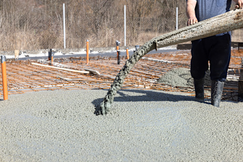 A worker guides the spout of a concrete mixer on a construction site as concrete pours out, forming a foundational slab.