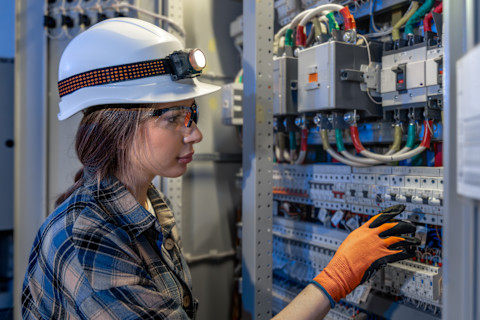 A worker wearing a helmet, frontal lamp, safety glasses and gloves reaches into an electrical panel.